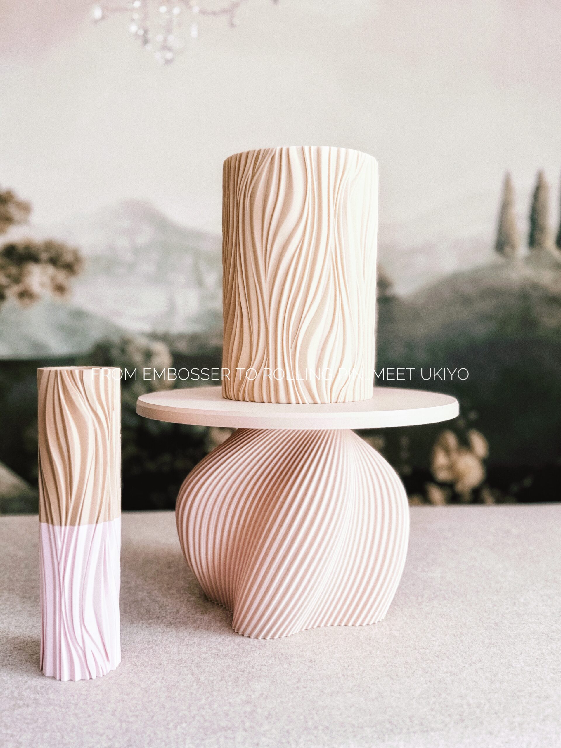 Ukiyo cake decorating texture shown as both an embosser and rolling pin. The design features elegant flowing wave-like lines that create a seamless, organic pattern on fondant-covered cakes. Displayed alongside a textured cake and sculptural cake stand in soft neutral tones, highlighting the modern, artistic finish achievable with the Ukiyo collection.